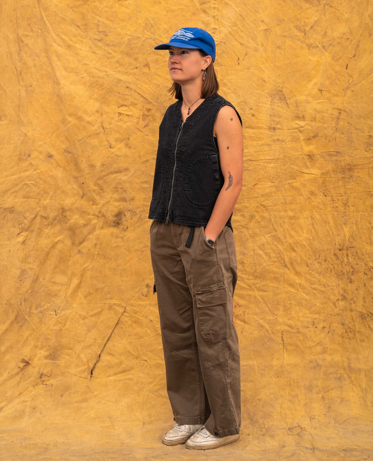 A woman stands in profile wearing a blue baseball cap, a black sleeveless zip-front vest, brown cargo pants, and white sneakers, posed against a textured yellow backdrop.