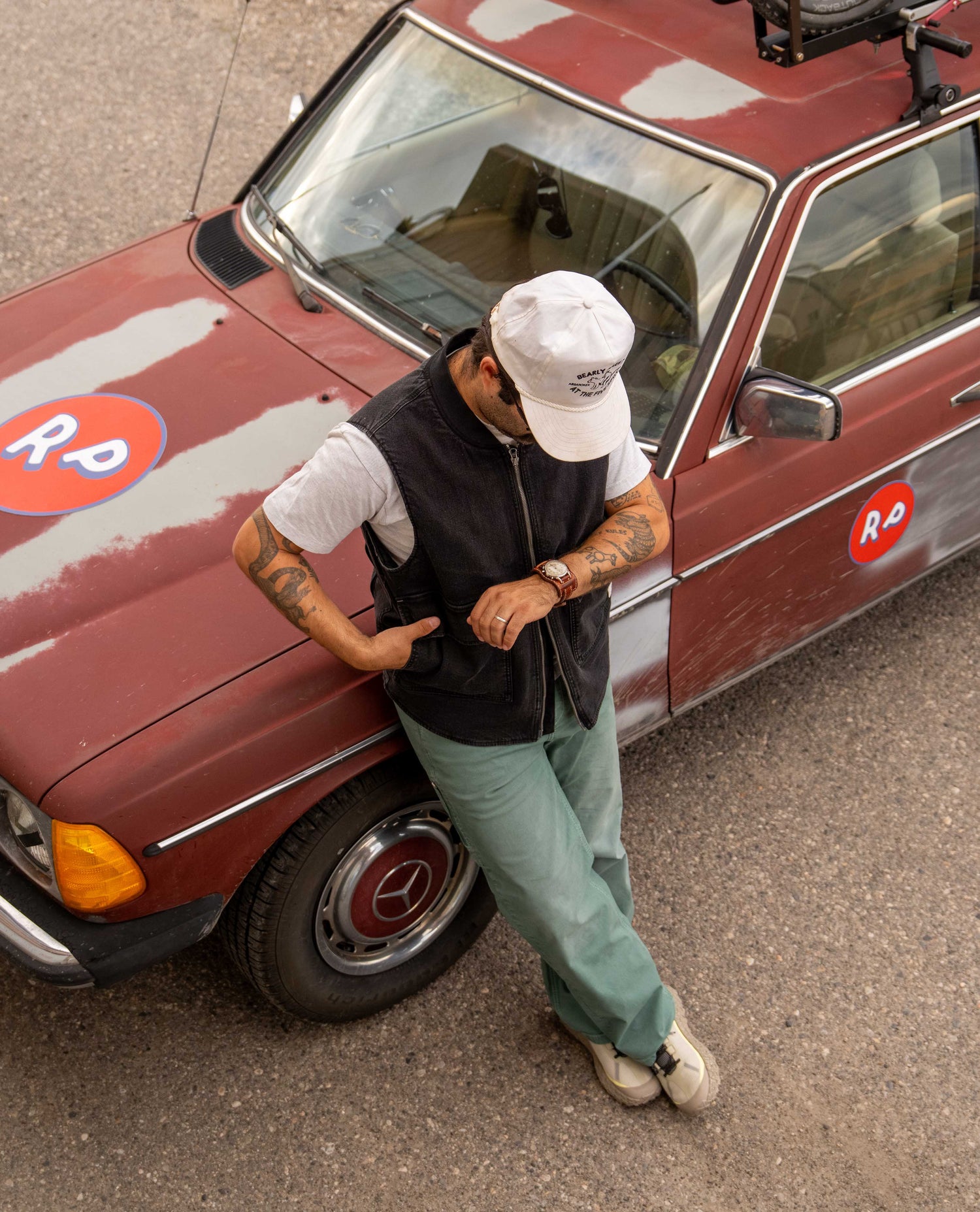 A man wearing a white baseball cap, black zip-up vest, white t-shirt, and light green pants leans against a vintage red car with a 'RD' logo, photographed from an overhead angle.