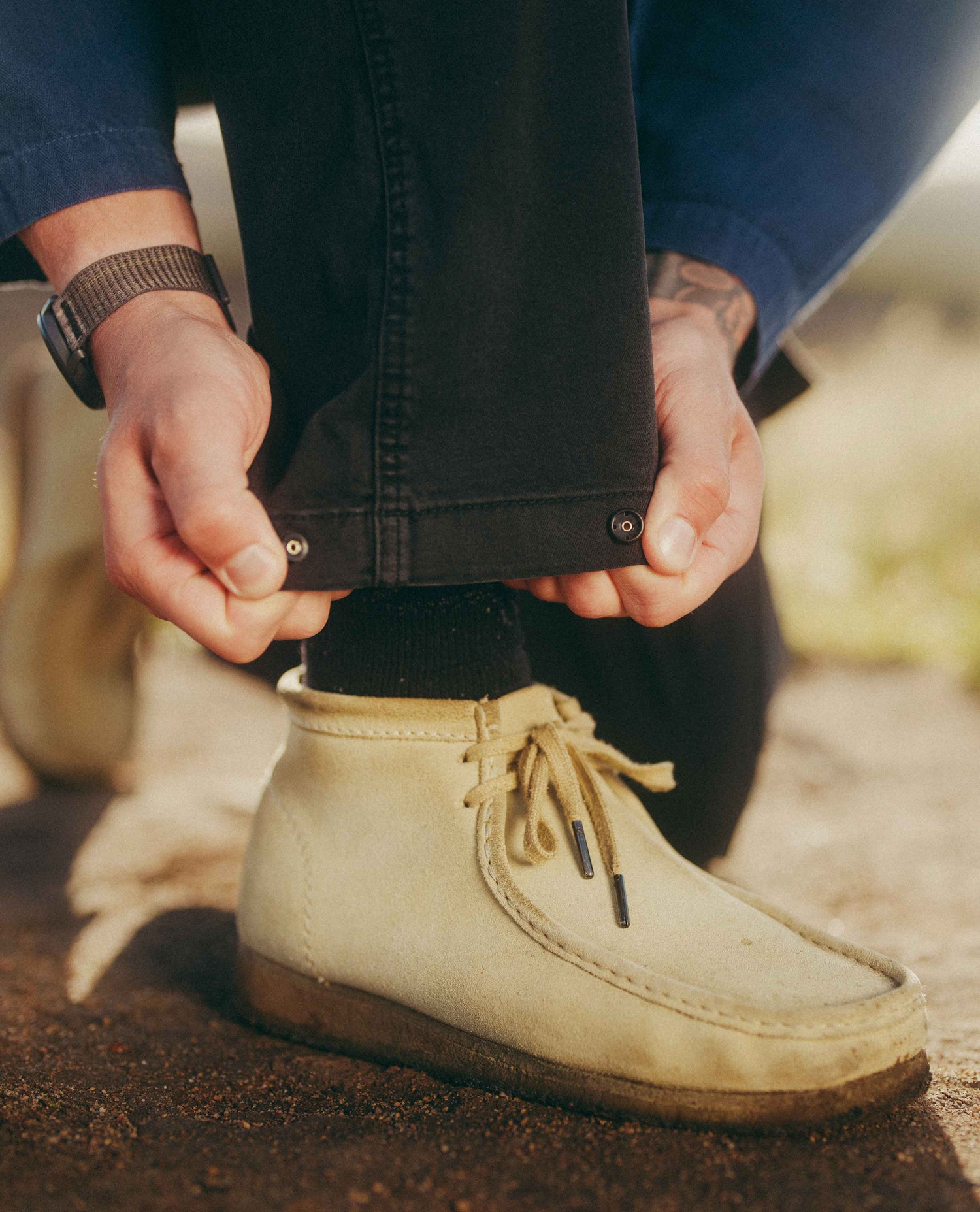 A close-up of a person's hands pulling down the hem of black jeans to reveal metal rivet tabs, wearing a beige suede mid-calf lace-up shoe and a dark sock.