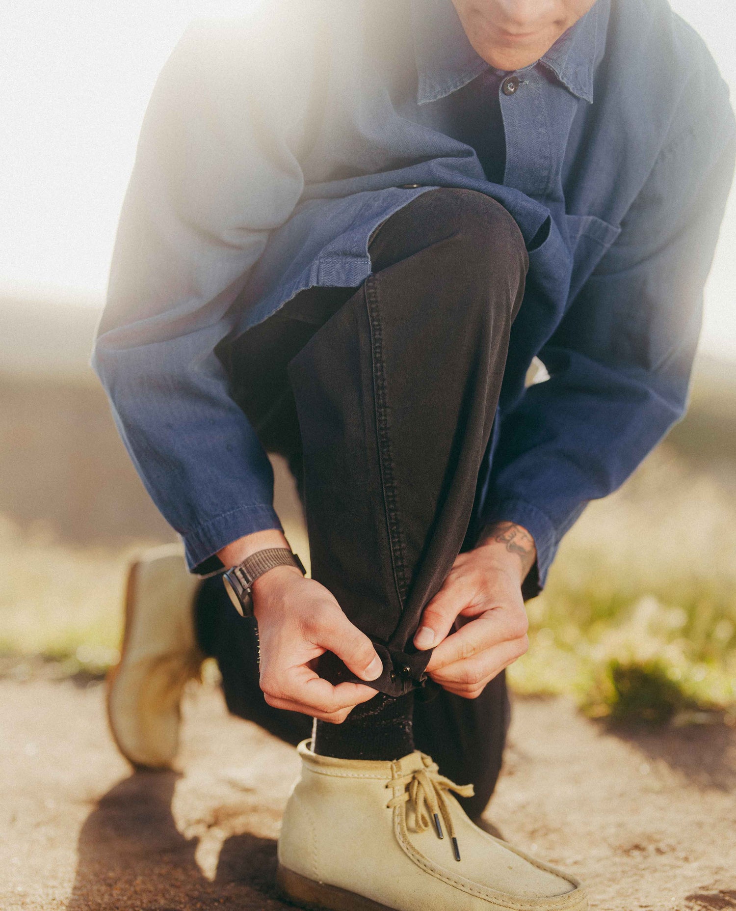 A man outdoors kneeling down to tie the laces of his black pants, wearing a blue button-up shirt and beige boots.