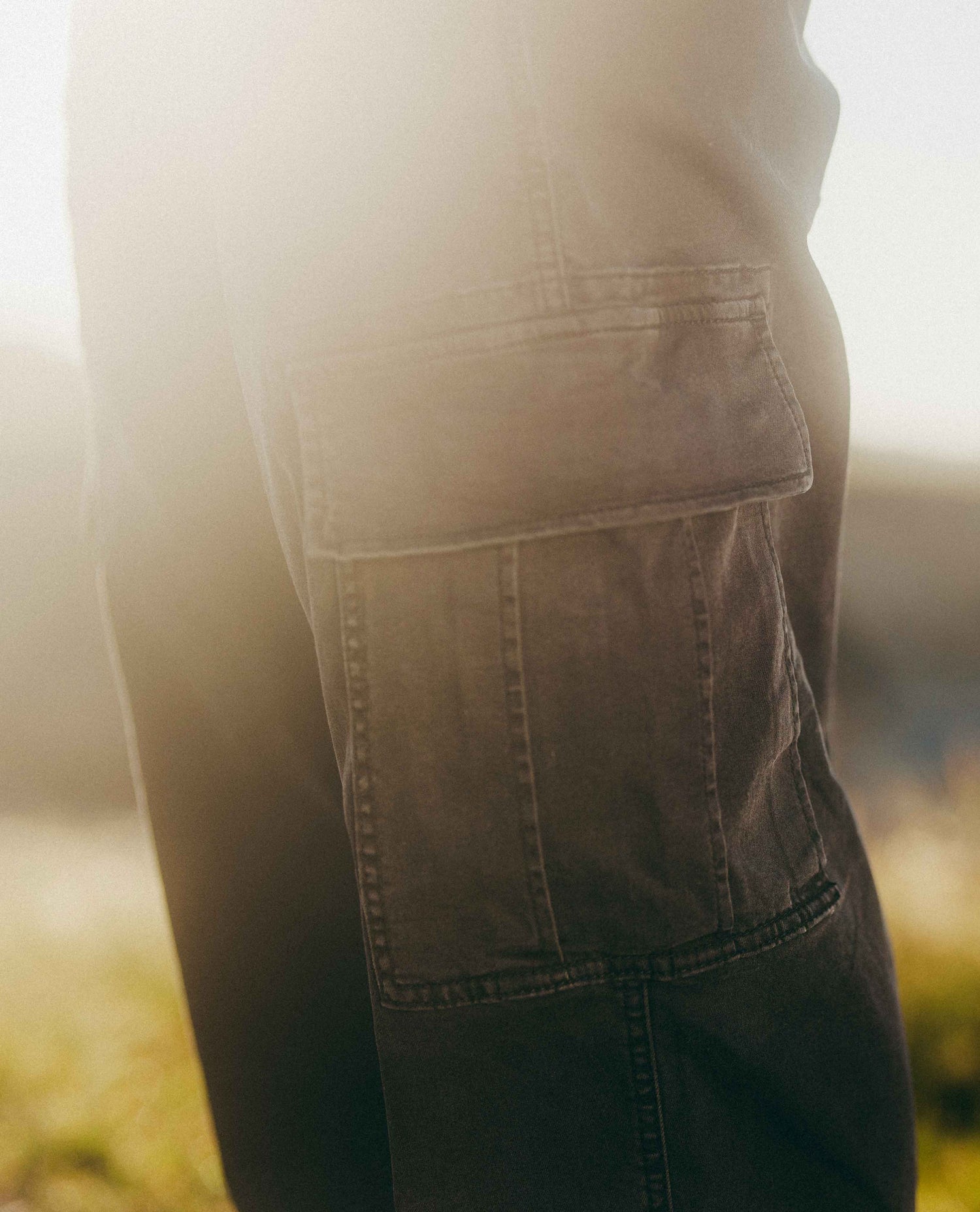 Close-up of dark gray cargo pants with a large flap pocket, shown outdoors in warm backlighting.