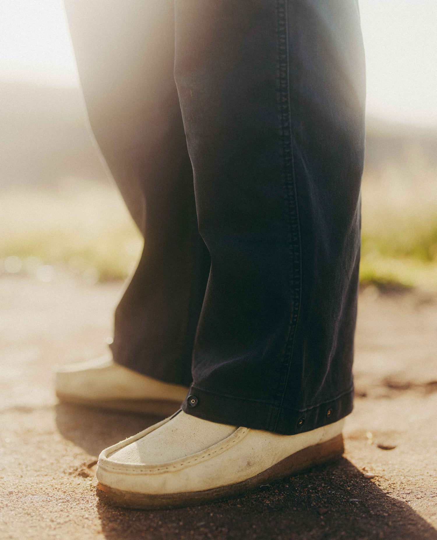 Close-up of a person's legs wearing dark navy blue jeans and off-white slip-on shoes, standing outdoors on a sunlit paved surface.