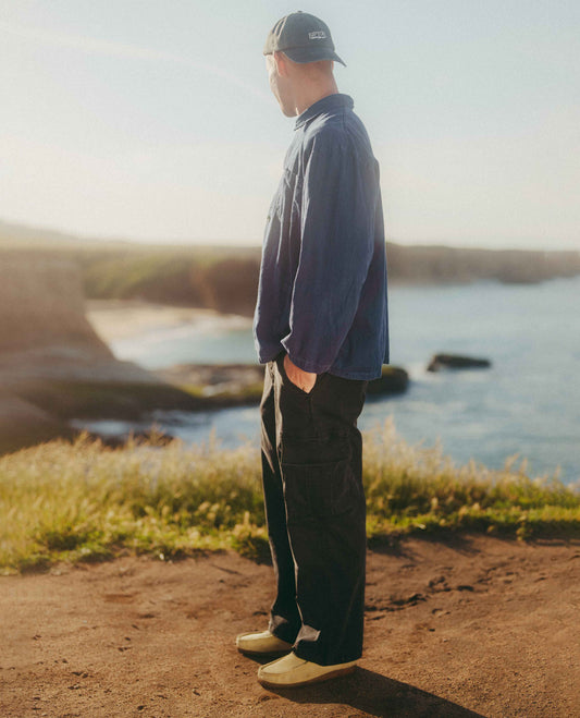 A man stands in profile outdoors by the water, wearing a dark navy overshirt, dark cargo pants, a dark baseball cap, and light-colored shoes. The background features a coastal landscape with rocks, grass, and a warm, sunlit sky.