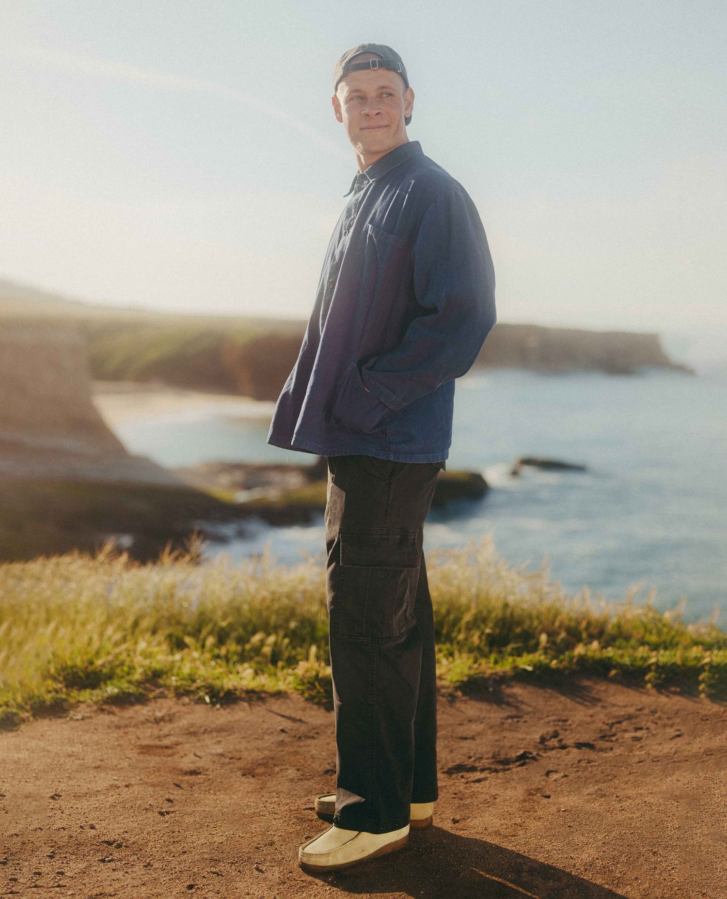 A man stands on a dirt path overlooking a rocky coastline at sunset, wearing a navy blue windbreaker jacket, dark cargo pants, and a dark baseball cap.