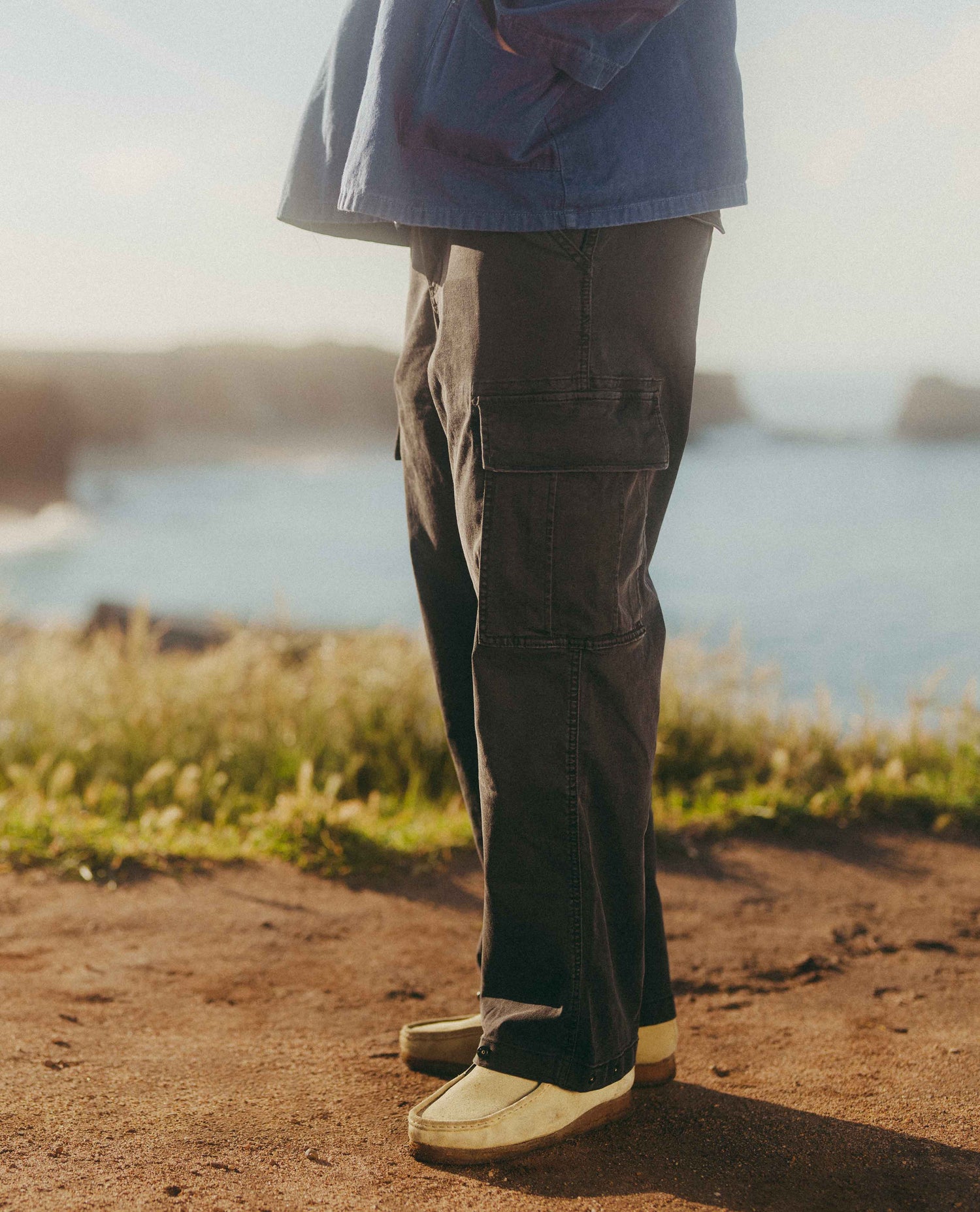 A person standing outdoors by the water wearing dark cargo pants, a blue shirt, and cream slip-on shoes. The photo is cropped from the chest down, with the pants and shoes most prominent against a warm coastal background.