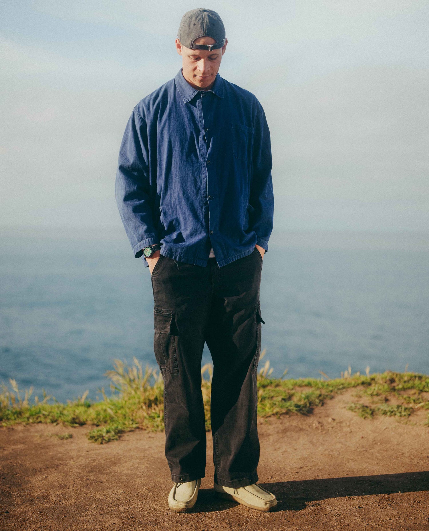 A man standing outdoors on a grassy cliff overlooking the ocean, wearing a navy blue button-up overshirt, black cargo pants, and a gray baseball cap.