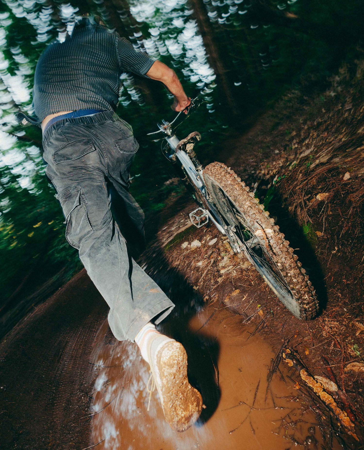A person riding a mountain bike through a muddy forest trail, viewed from behind and below. The rider wears a dark patterned shirt, dark pants, and white socks, with the bike's front wheel and muddy tire visible.
