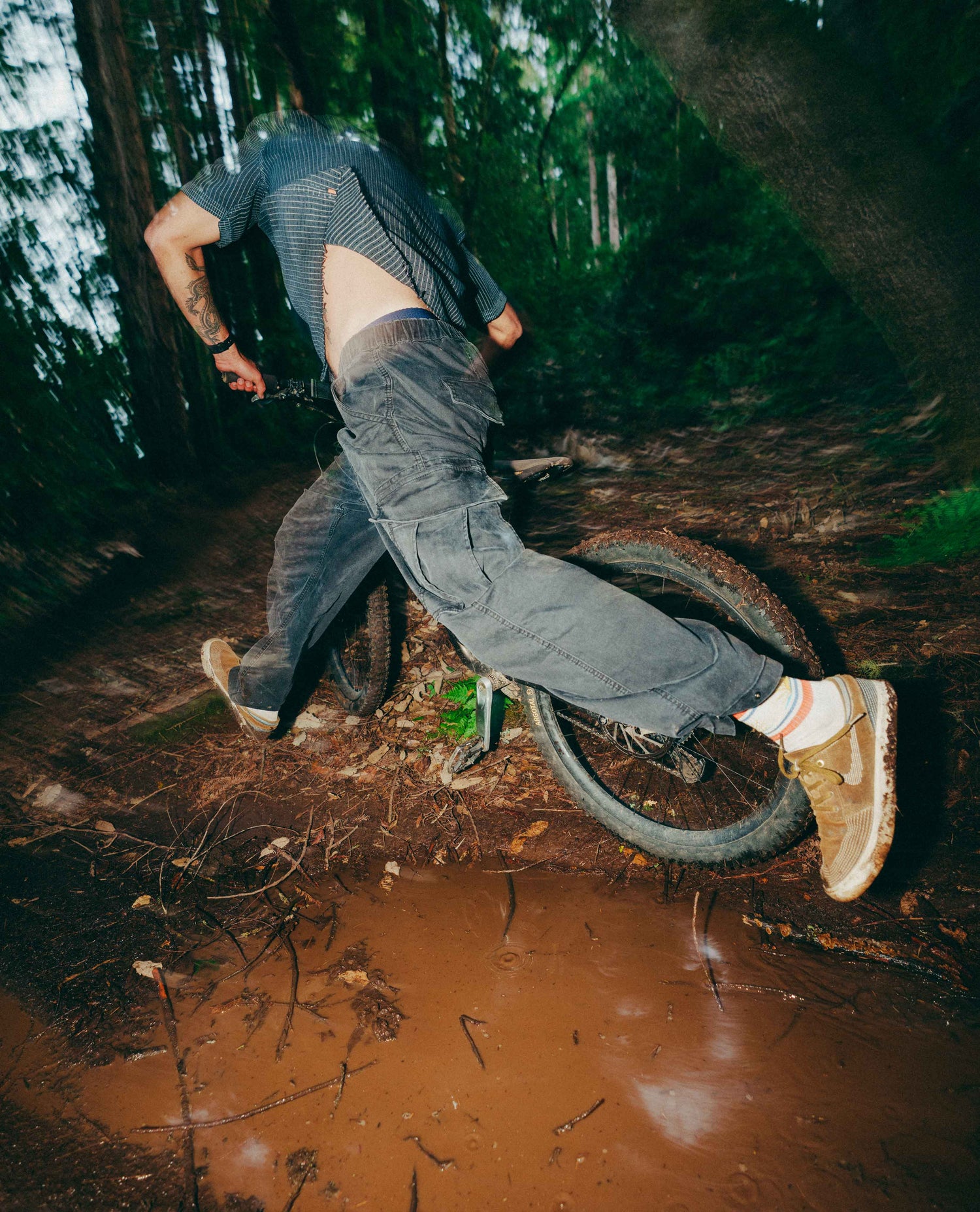 A person riding a mountain bike through a muddy forest trail, viewed from behind. The rider wears a dark striped short-sleeve shirt, loose gray cargo pants, white socks, and tan sneakers.