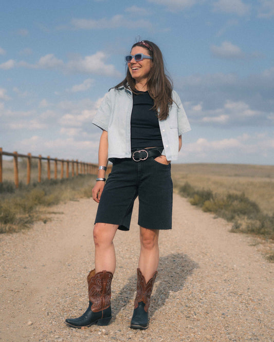 A woman stands on a dirt path outdoors wearing a light blue short-sleeve button-up shirt over a black top, black shorts, a black belt with a silver buckle, and dark cowboy boots with brown shaft detailing. She is wearing sunglasses, and the background shows a grassy field under a blue sky with clouds.