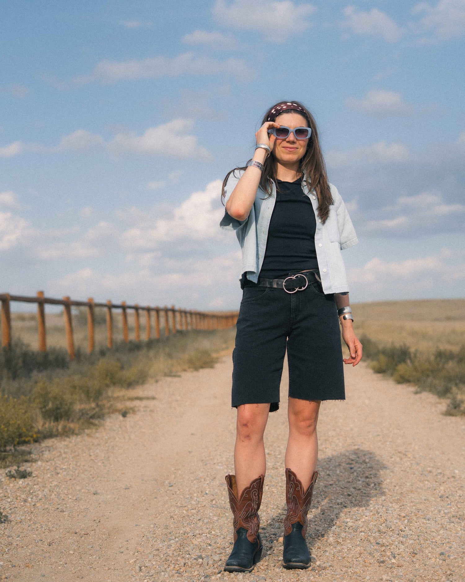 A woman stands on a dirt path outdoors wearing a light blue denim shirt, black top and shorts, a black belt with a silver buckle, brown cowboy boots, and sunglasses.