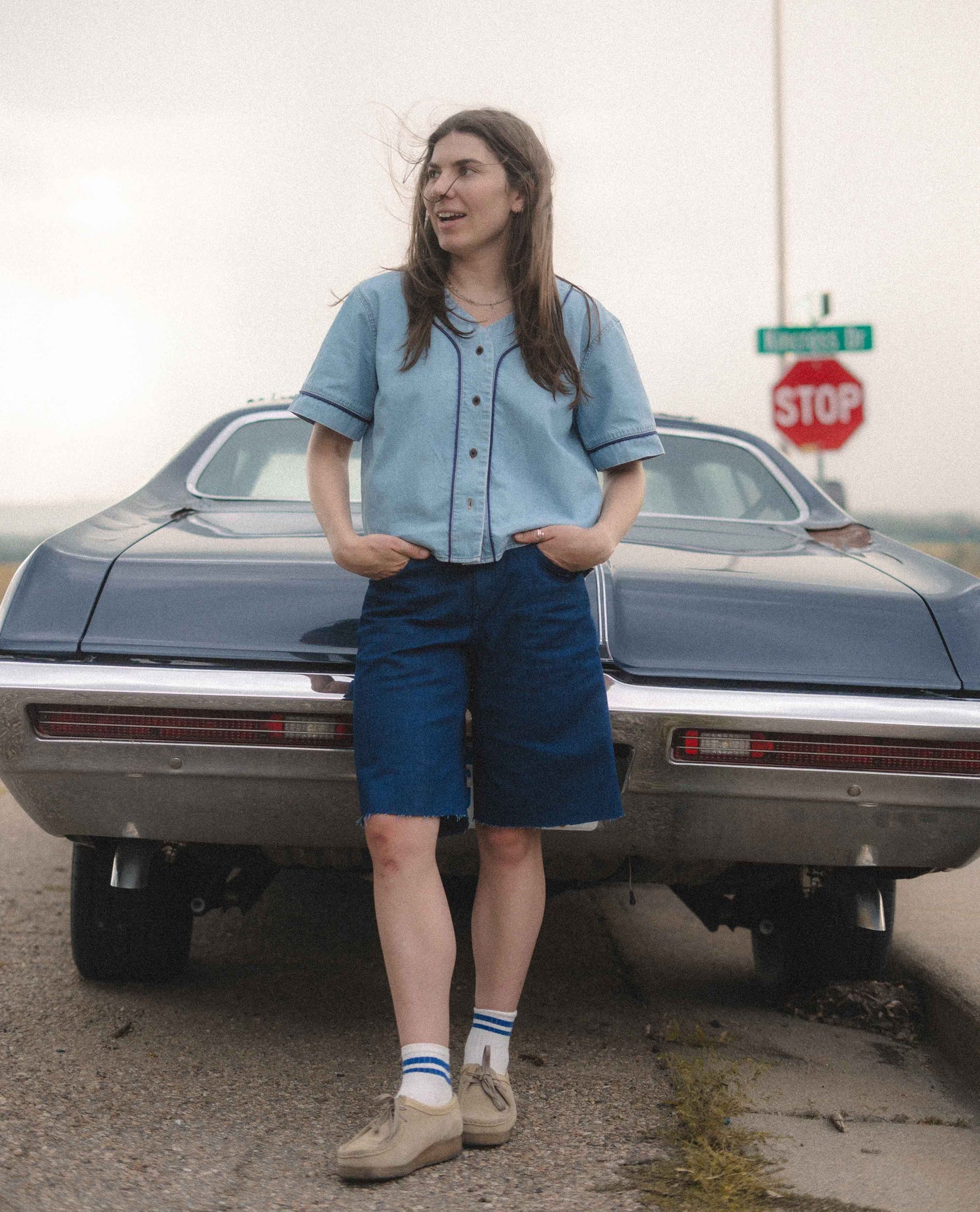 A person stands in front of the rear of a dark blue classic car on an outdoor road, wearing a light blue short-sleeve button-front shirt, dark blue shorts, white socks, and tan shoes.