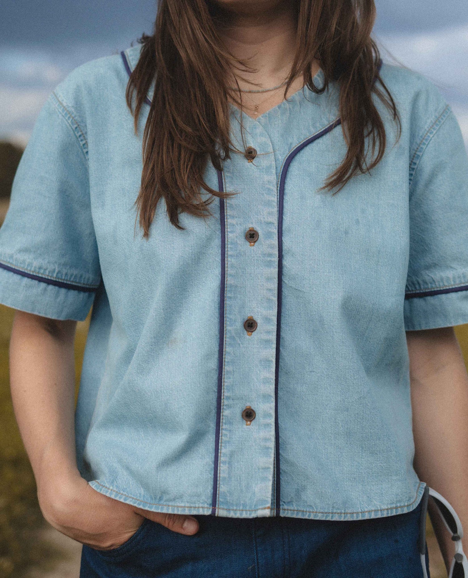 A person wearing a light blue denim short-sleeve button-front shirt with dark contrast piping and brown buttons, paired with dark blue jeans, photographed outdoors against a blurred sky and grass background.