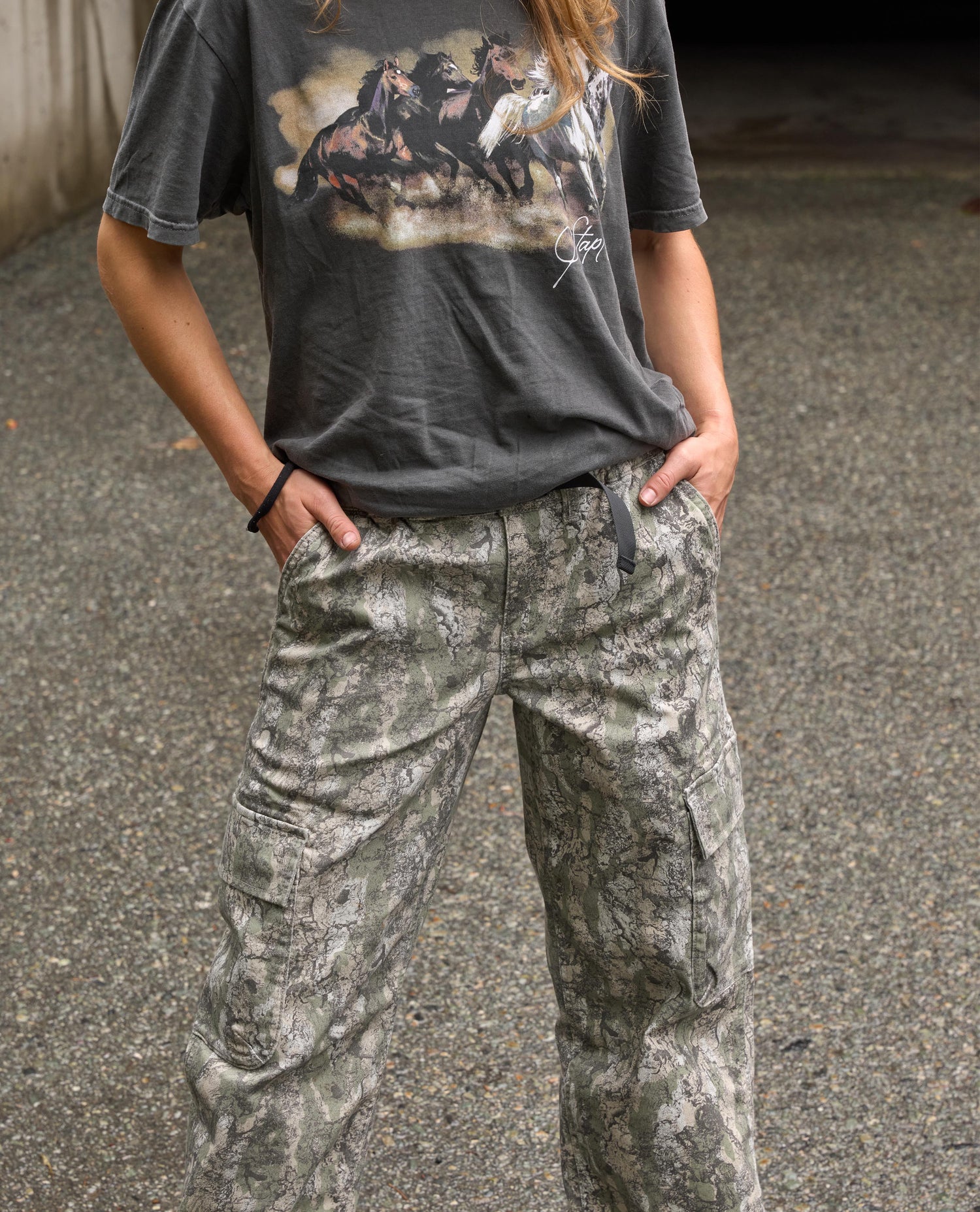 A person wearing a dark gray graphic t-shirt and wide-leg camouflage cargo pants, standing outdoors on a paved surface.