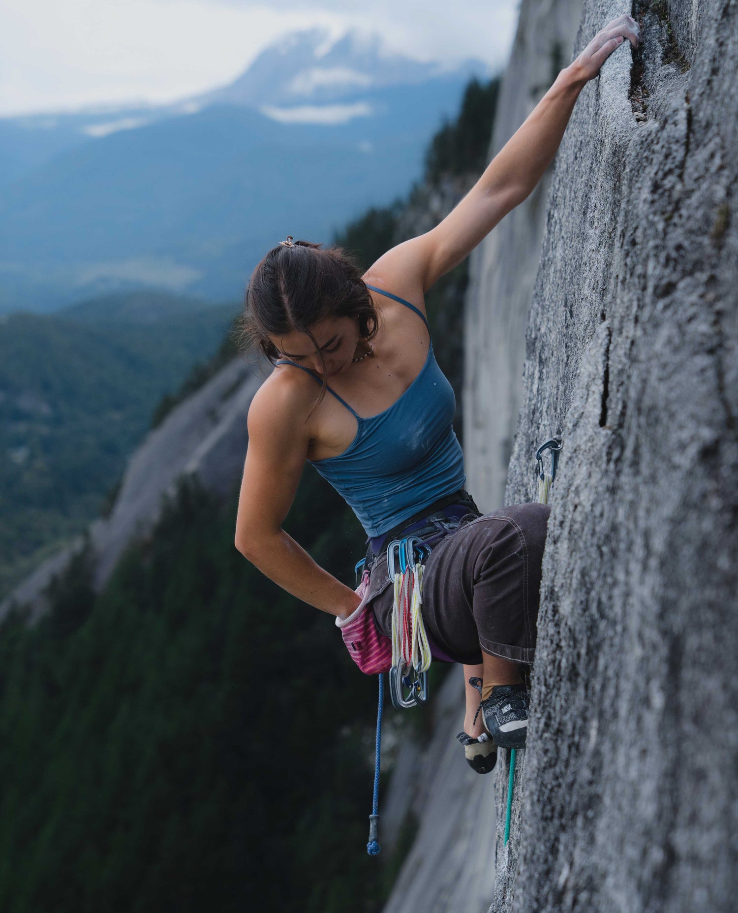 A woman climbing a rock wall reaching for her chalk bag wearing a blue top and brown pants