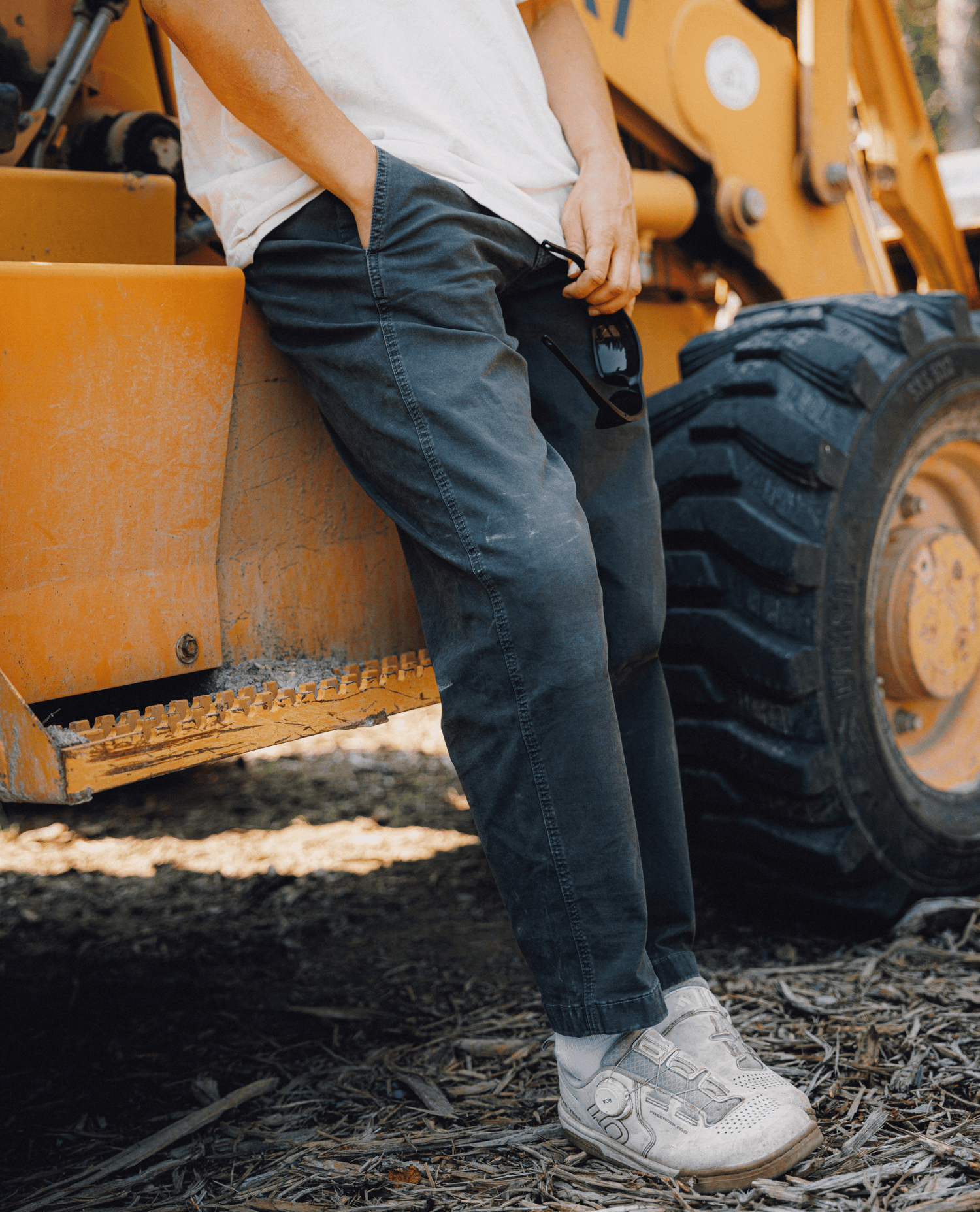 A person wearing dark charcoal gray pants and white sneakers leans against a yellow construction vehicle, with a large tire visible in the background.