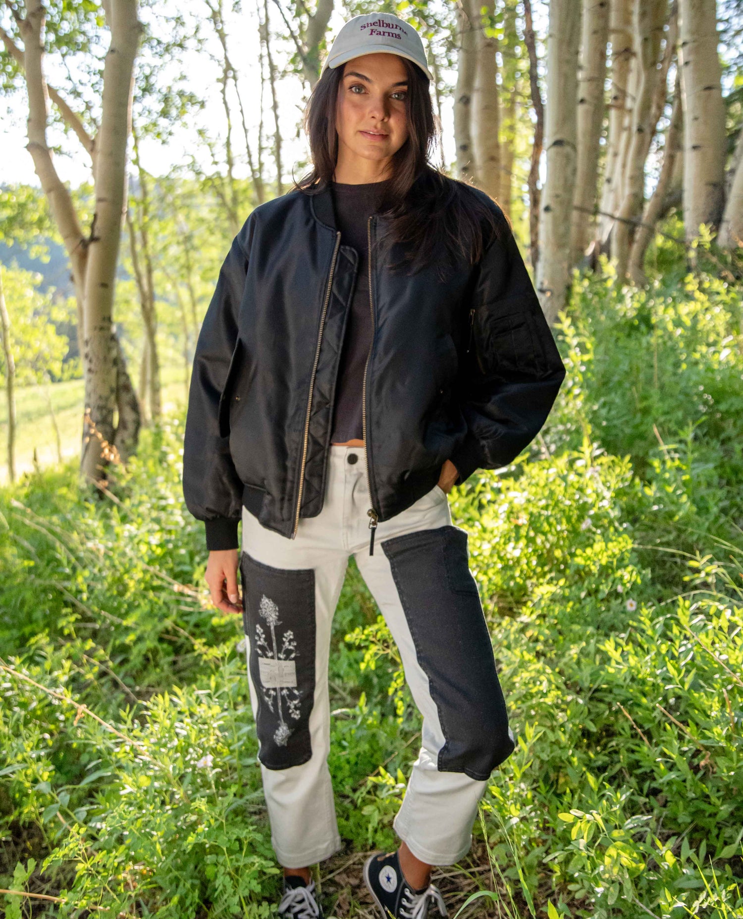 A woman stands outdoors in a wooded area wearing a black bomber jacket, a black t-shirt, white and black patchwork cargo pants, and a white baseball cap.