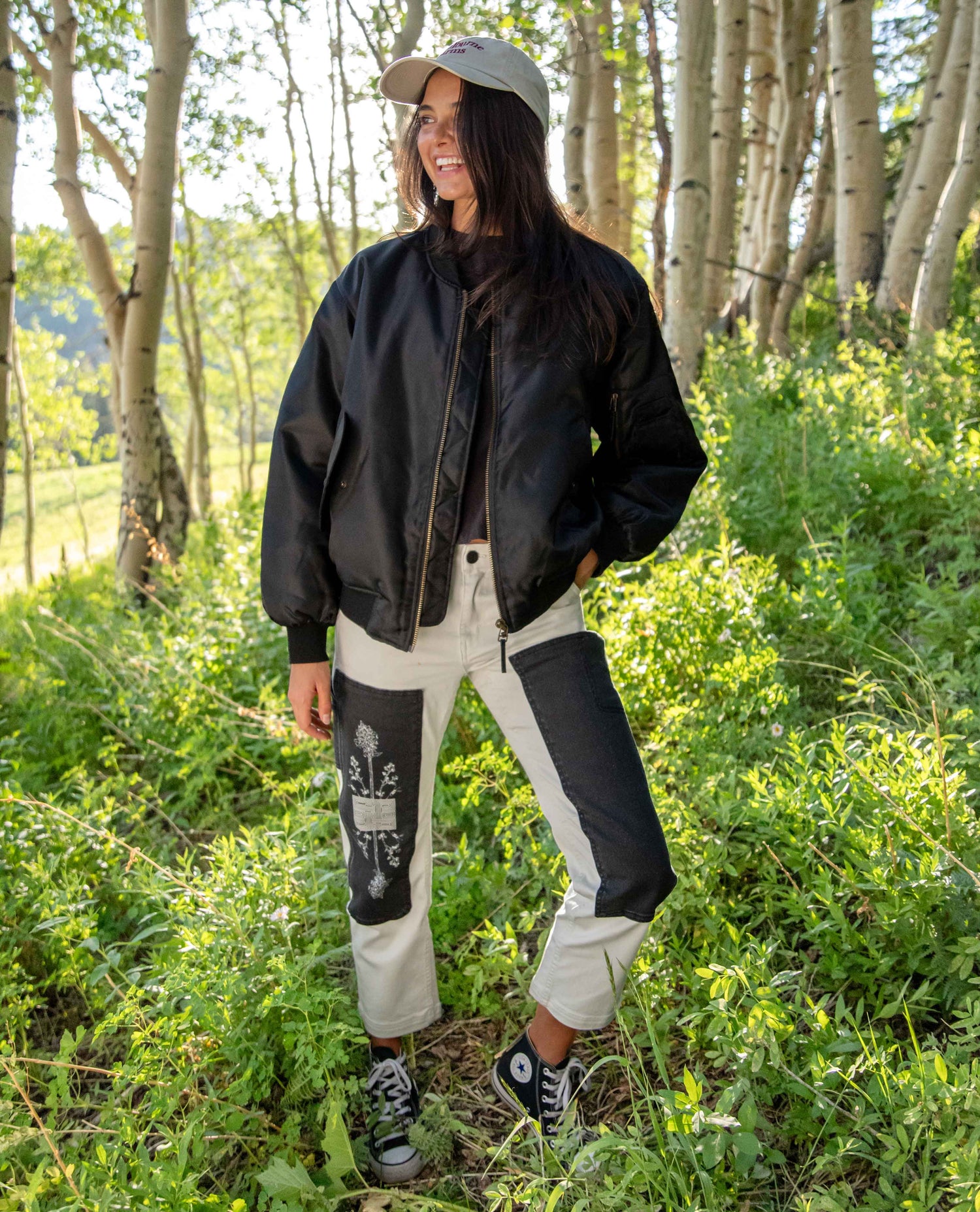 A woman stands outdoors in a wooded area wearing a black bomber jacket, off-white patchwork pants with black panels, a gray cap, and black-and-white sneakers.