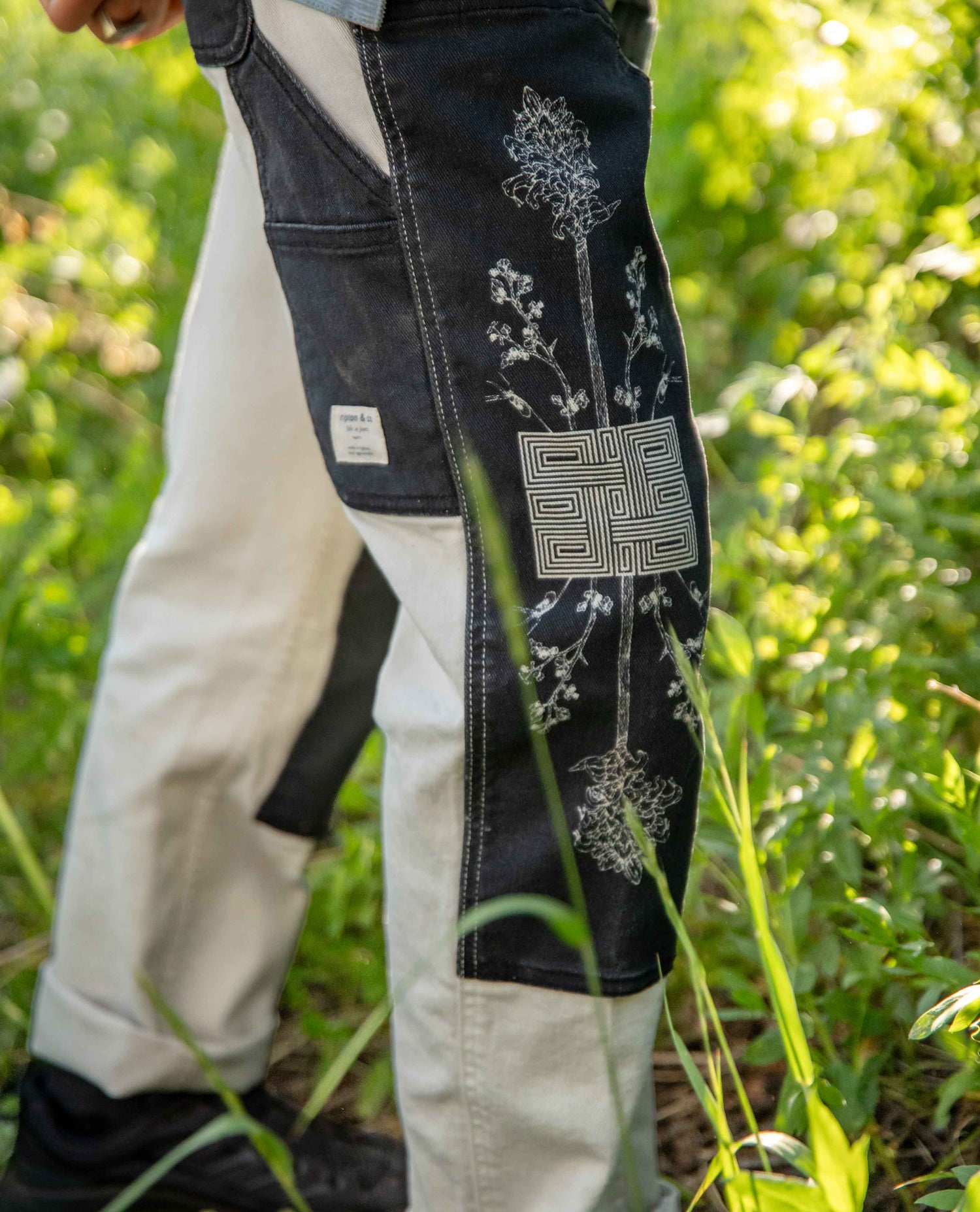 Close-up of a person outdoors wearing dark denim overalls with white embroidered floral and geometric patterns, paired with off-white pants and black shoes, set against a blurred green grassy background.