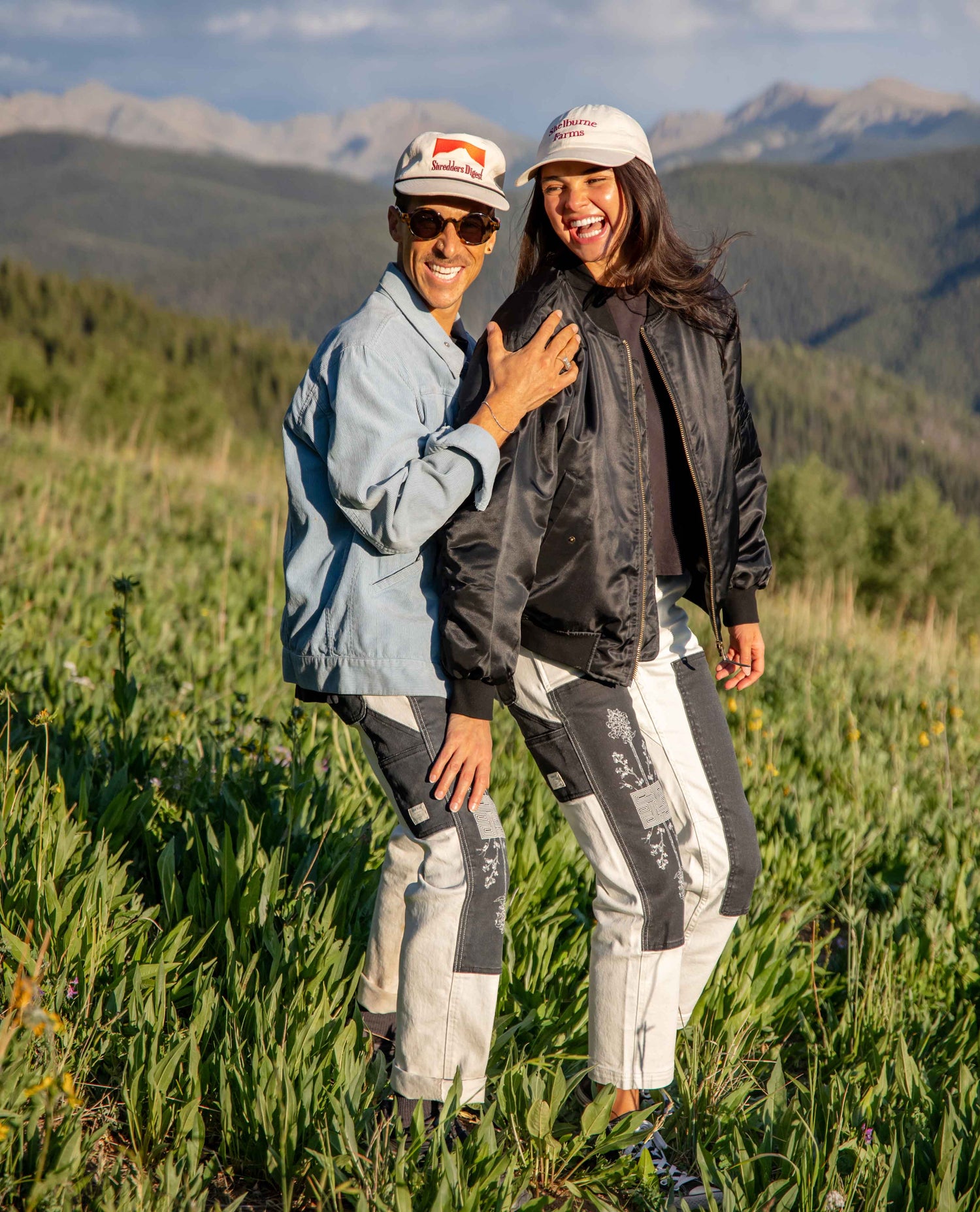 Two people standing outdoors in a grassy mountain landscape, wearing matching light-colored outdoor jackets and pants with dark gray paneling, plus white caps. The person on the right wears a black jacket, and the person on the left wears a light blue jacket.