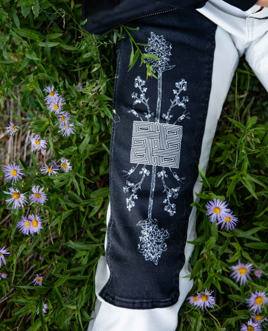 A close-up of a person's leg wearing black and white side-panel pants, lying on green grass with small purple flowers. The black fabric has a large white embroidered floral and geometric design on the outer thigh.