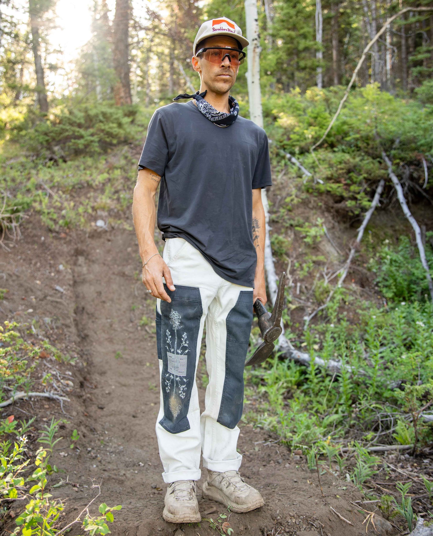 A man stands on a dirt trail in a forest wearing a dark gray t-shirt, off-white pants with dark gray patches, a bandana, sunglasses, and a cap.