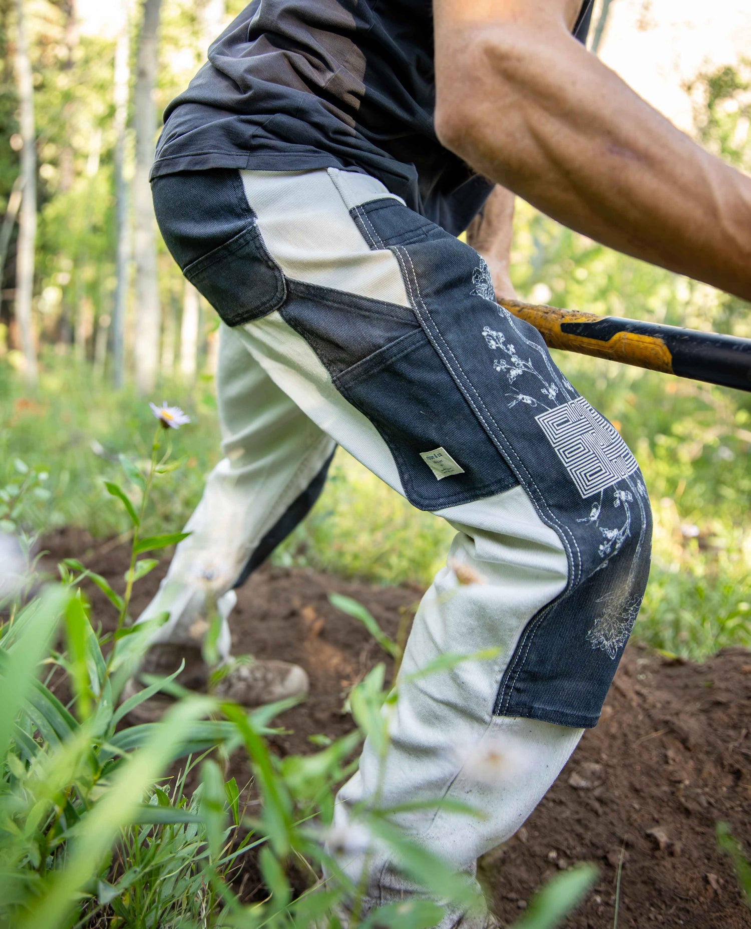 A person outdoors in a field wearing heavy-duty work pants with dark blue denim panels and off-white canvas sections, holding a shovel.