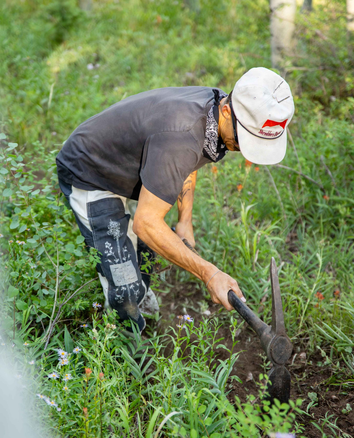 A man in a black t-shirt, white cap, and work pants bends over using a hand tool in a grassy outdoor field.