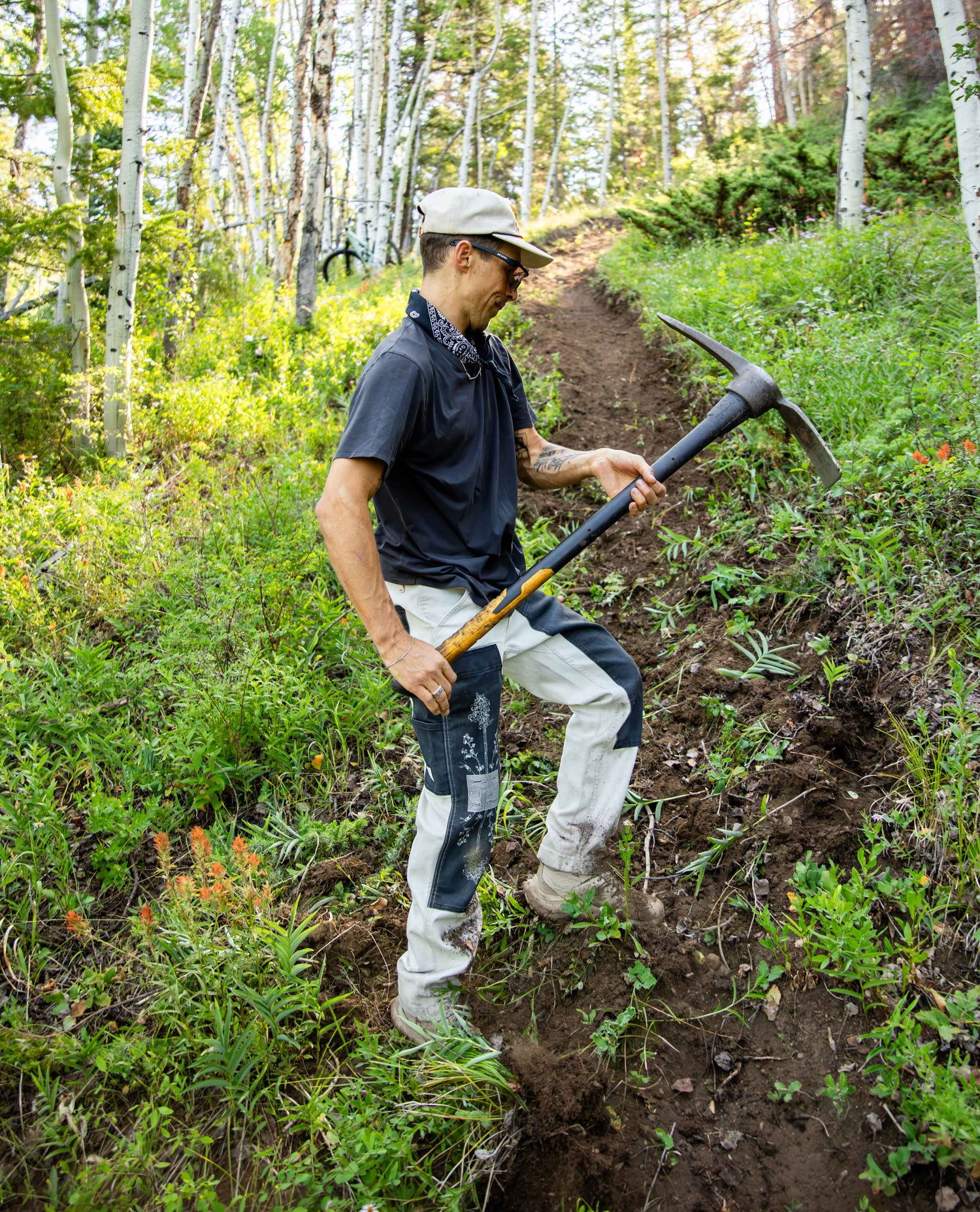A man wearing a beige cap, dark navy polo shirt, and off-white work pants with dark patches is shown in a forest clearing, holding a pickaxe and digging along a narrow path.