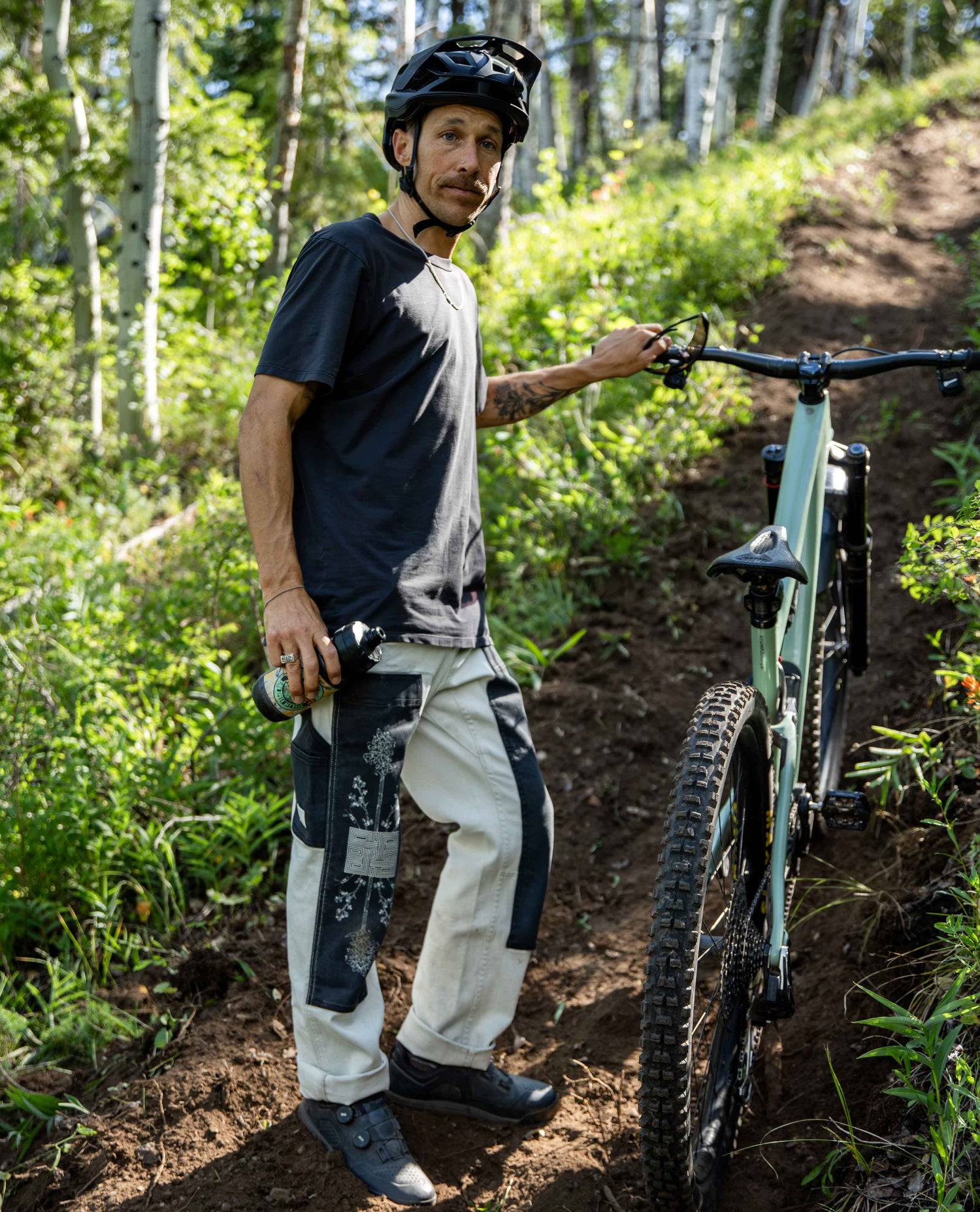 A mountain biker stands on a dirt trail in a forest, wearing a black helmet, a dark graphic t-shirt, and off-white pants with black knee patches, next to a green full-suspension mountain bike.