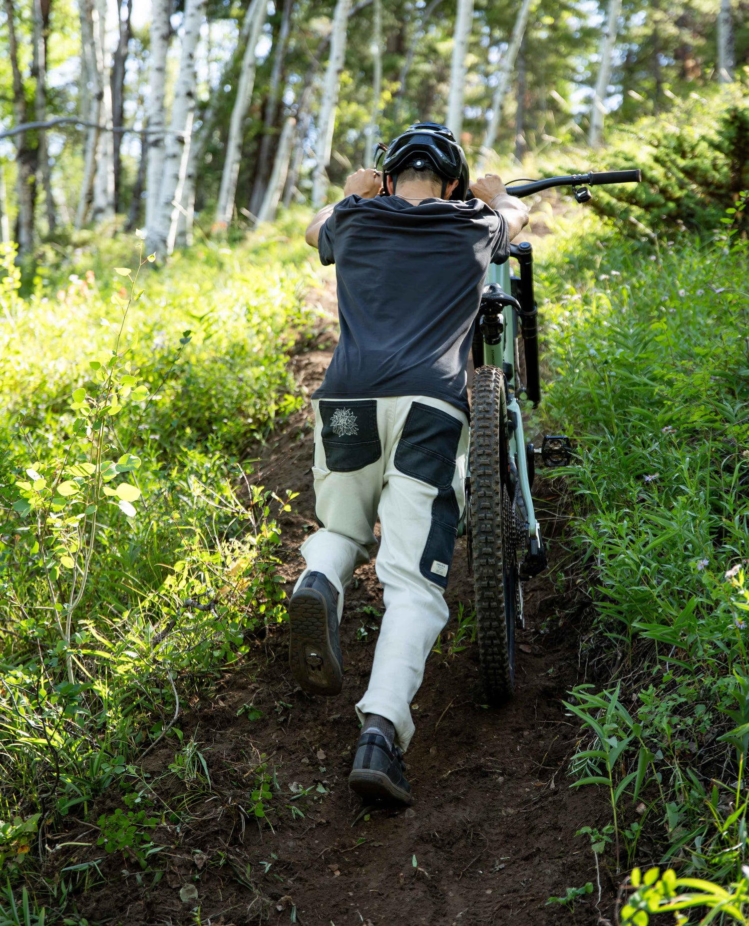 A person wearing a black helmet, dark gray t-shirt, and off-white technical mountain biking pants pushes a green and black mountain bike up a dirt trail in a lush green forest.