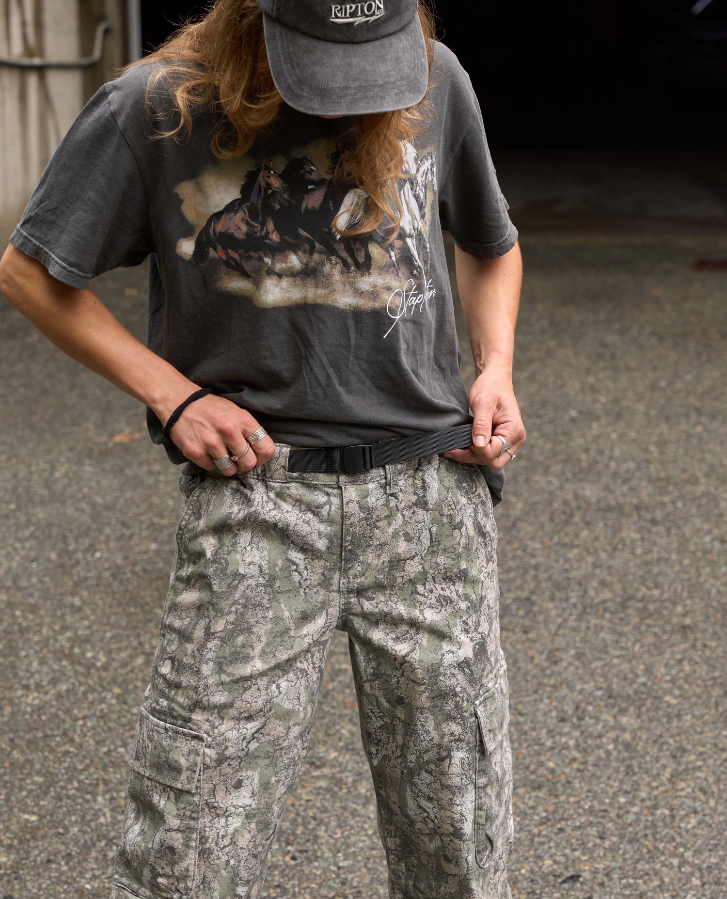 A person wearing a dark gray graphic t-shirt, a black cap, and digital camouflage cargo pants, standing outdoors on asphalt.