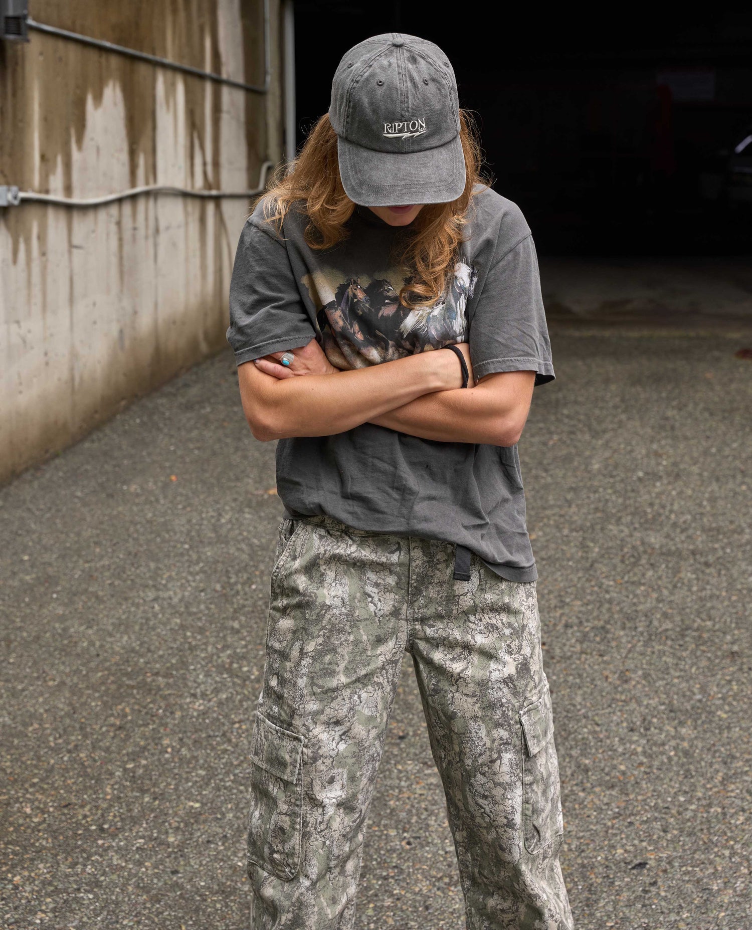 A person standing outdoors with arms crossed, wearing a dark gray graphic t-shirt, camouflage cargo pants, and a gray baseball cap with a visible 'Kipsta' logo.