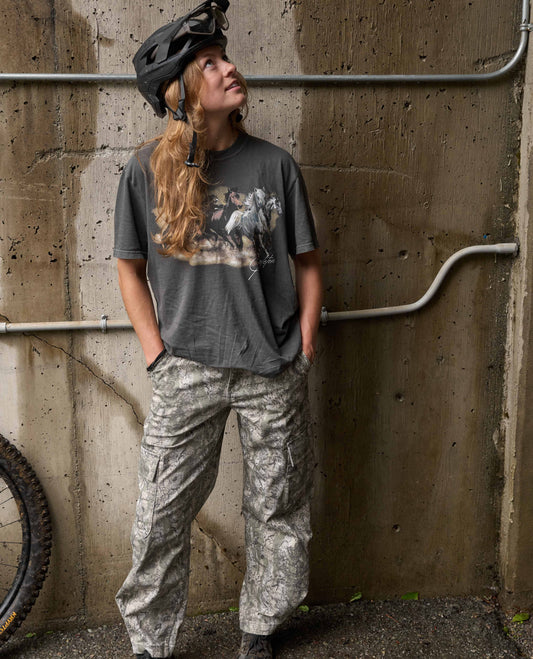 A woman wearing a dark gray graphic t-shirt, camouflage cargo pants, and a black bicycle helmet stands against a concrete wall.