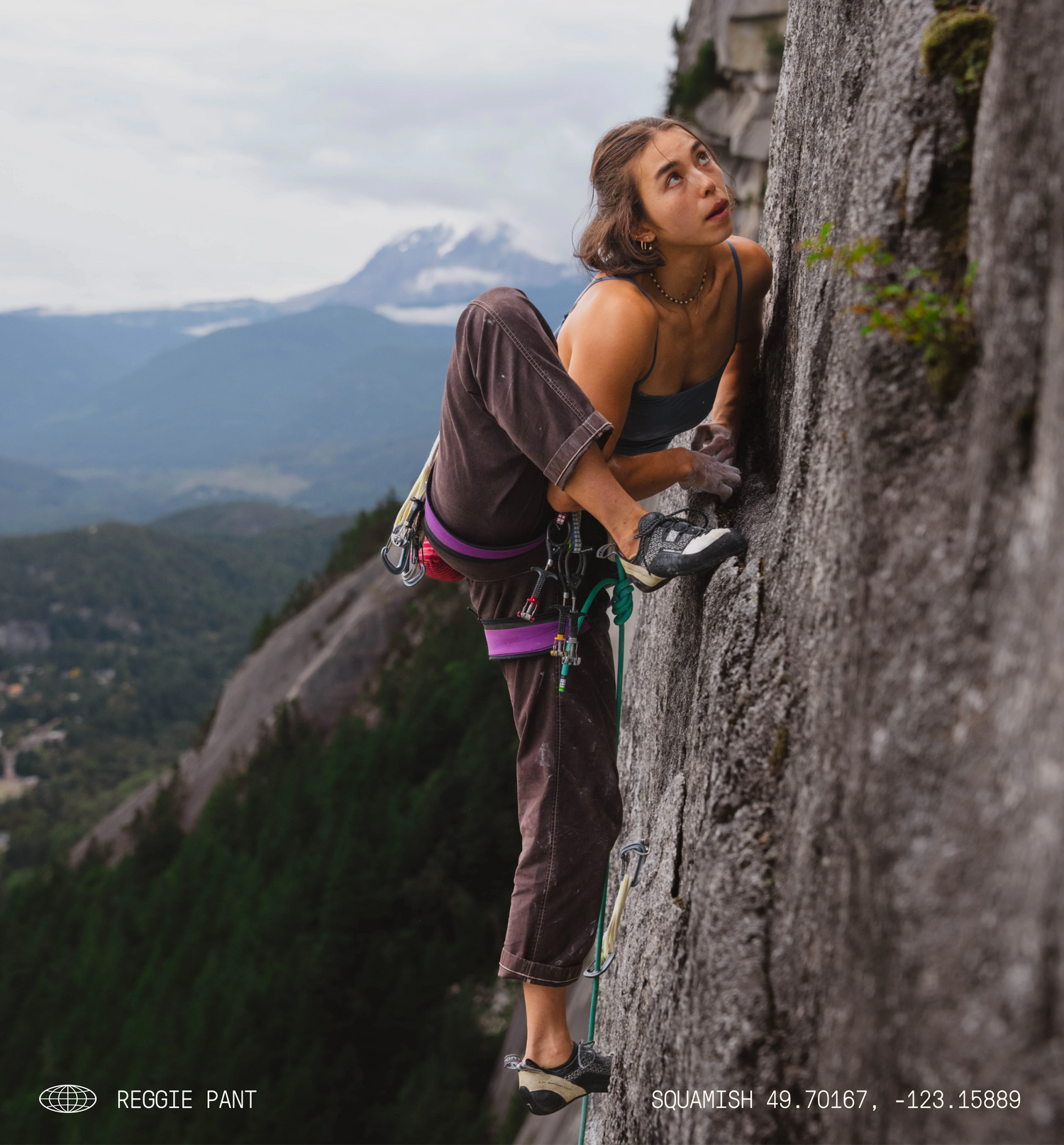 A climber in brown pants ascends a granite rock face, with a mountain landscape in the background. Text at the bottom reads “REGGIE PANT” and “SQUAMISH 49.70167, -123.15889.”