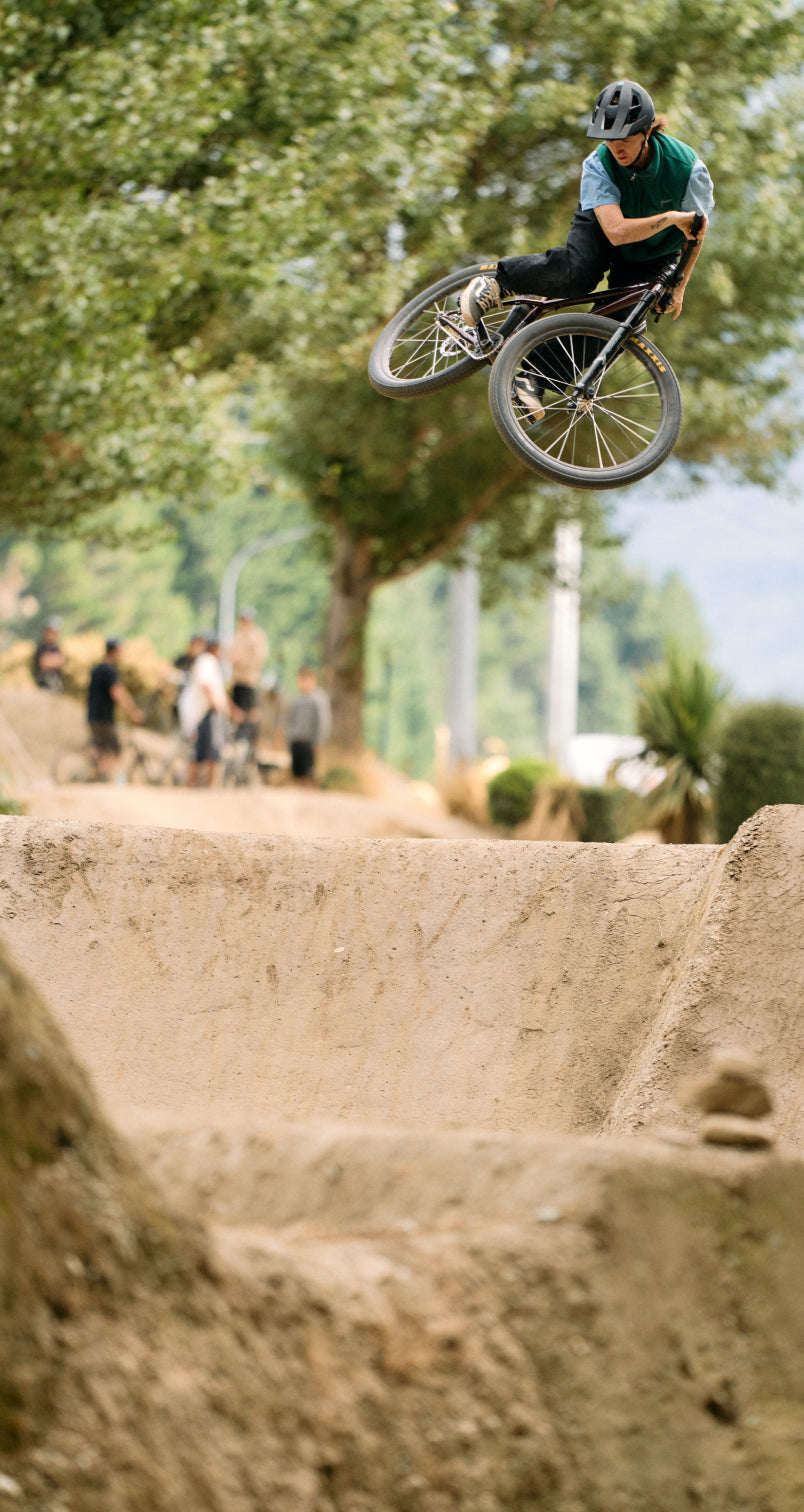 Person on a bicycle performing a jump in an outdoor setting with trees and people in the background.