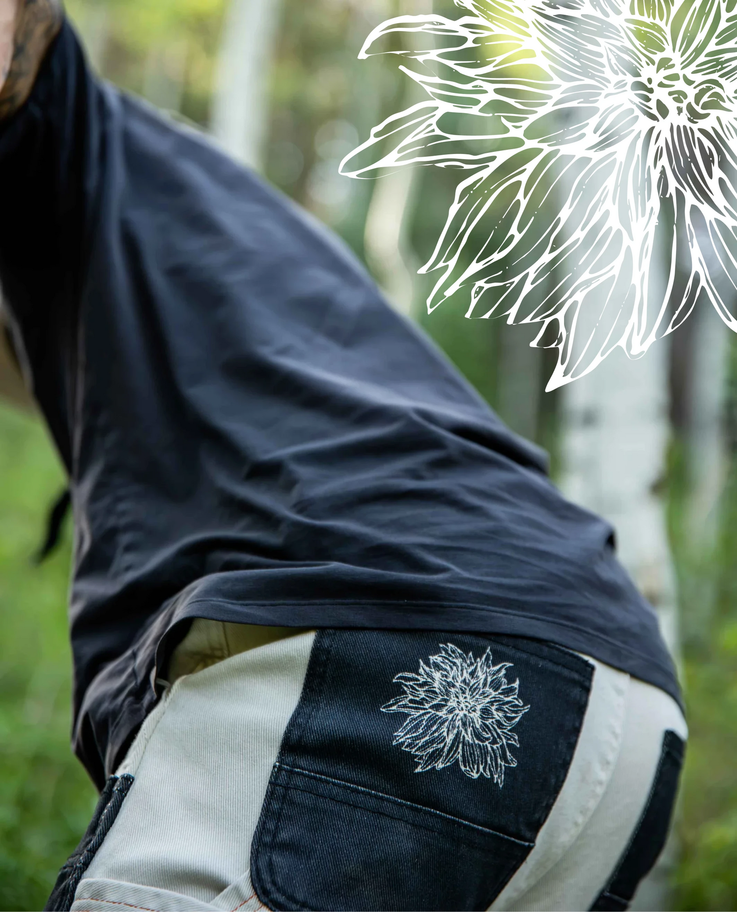 A close-up outdoor shot of a person wearing a dark navy T-shirt and light beige shorts with a white floral line-art graphic printed on the back pocket. The background is a soft-focus green forest.