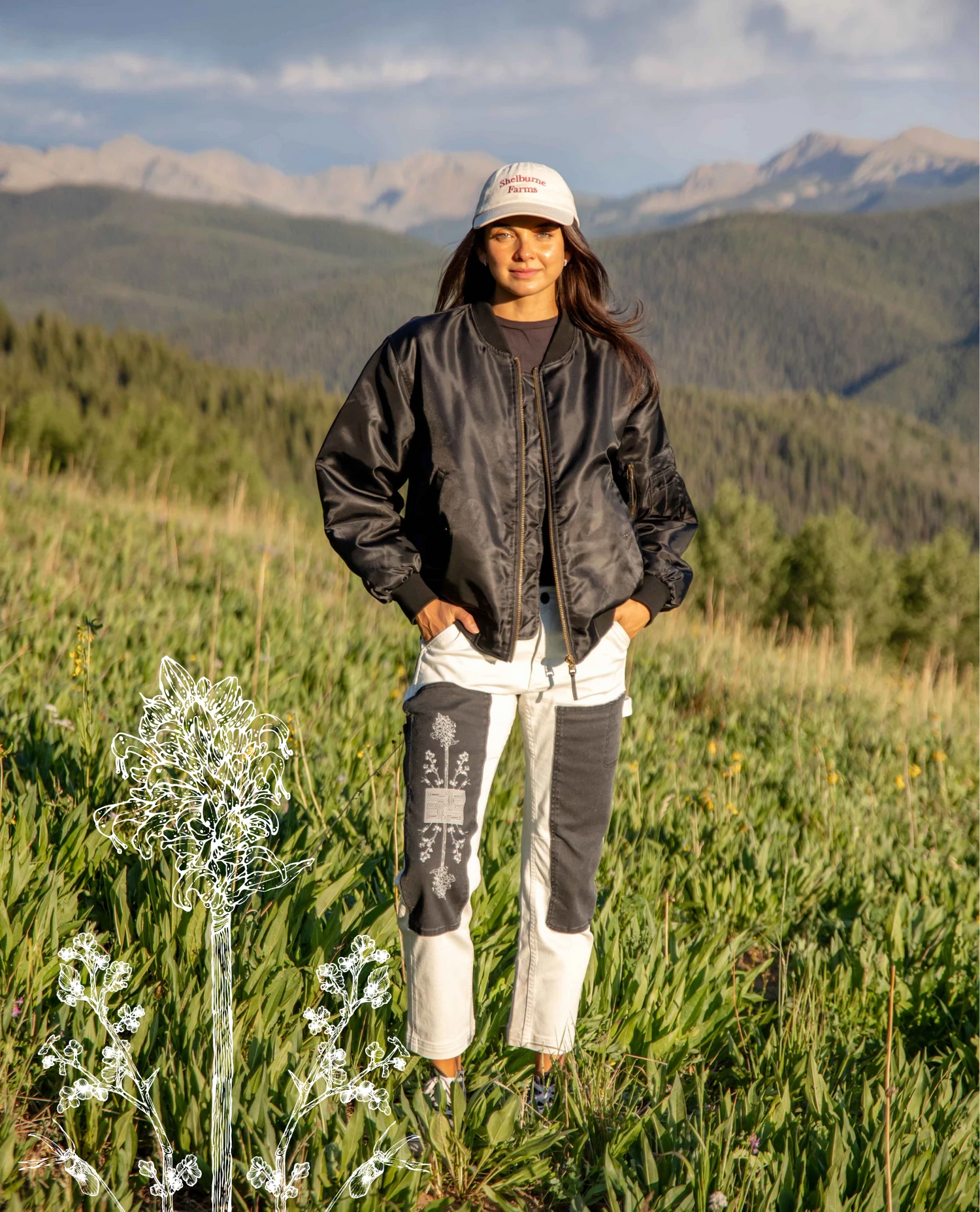 A woman stands in a grassy mountain landscape wearing a black bomber jacket, white and black patchwork pants, a white cap, and dark boots. The background features green hills and distant mountains under a cloudy sky.
